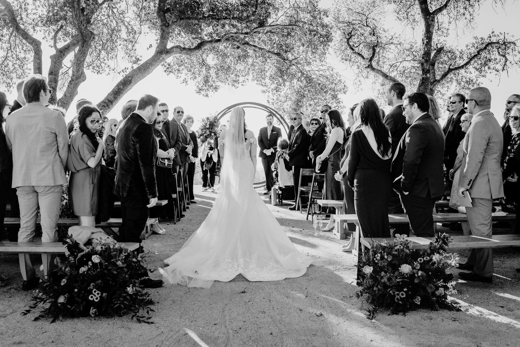 A bride walking down an outdoor aisle under old oak trees toward the arch