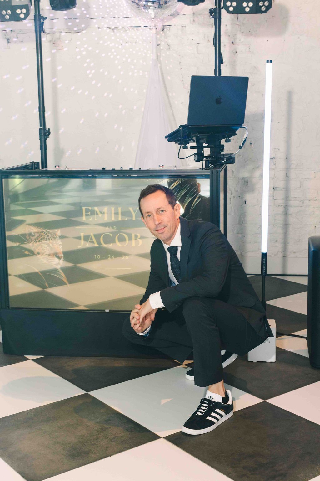 Matt crouching on a checkered dance floor in a suit and sneakers, smiling beside his booth